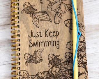 May include: A wooden spiral bound journal with a gold coil. The cover is engraved with a floral design and goldfish. The text "Just Keep Swimming" is engraved on the cover. A yellow pencil is attached to the journal with a blue rubber band.