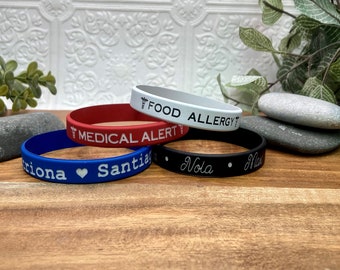 May include: Four silicone wristbands with different text and colors. The top band is white with black text that reads "FOOD ALLERGY". The second band is red with white text that reads "MEDICAL ALERT". The third band is blue with white text that reads "Triona Santi". The fourth band is black with white text that reads "Nola Mas".