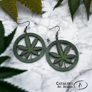 May include: A pair of green wooden earrings featuring a cannabis leaf design within a circular frame. The earrings have black hooks and are set against a marble background. The text "CATALYST Art. Design." is visible.