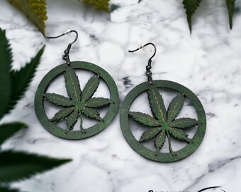 May include: A pair of green wooden earrings featuring a cannabis leaf design within a circular frame. The earrings have black hooks and are set against a marble background. The text "CATALYST Art. Design." is visible.