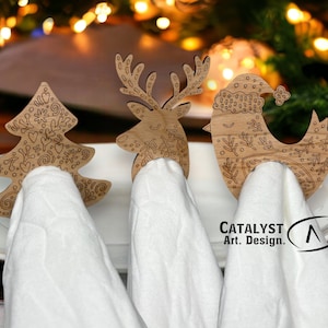 May include: Three wooden napkin rings with a Christmas theme. The rings are shaped like a reindeer, a Christmas tree, and a bird wearing a Santa hat. The rings are on white napkins and a white plate. The text "Catalyst Art. Design." is visible in the bottom right corner of the image.