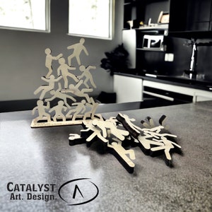 May include: A wooden game featuring a stack of stick figures, with additional figures scattered on a gray surface. The figures are all in the same pose, with arms outstretched and legs spread apart. The game is called "Catalyst Art. Design."