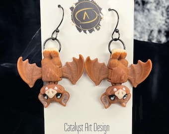 May include: A pair of brown bat earrings with black hooks. The bats are hanging upside down with their wings spread out. The earrings are made of polymer clay and have a cute, cartoonish look. The earrings are on a white card with the text "Catalyst Art Design" and "Rochester, MN".