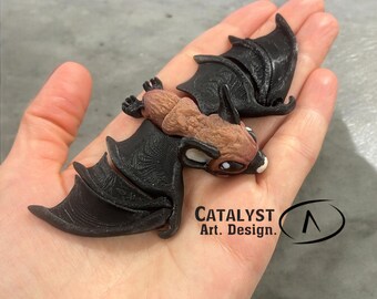 May include: A 3D printed brown and black bat figurine with a detailed texture. The bat is posed with its wings spread out. The figurine is held in a person's hand. The text "Catalyst Art. Design." is visible in the image.