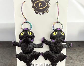 May include: A pair of black cat dangle earrings with purple accents and iridescent rainbow hooks. The earrings are displayed on a white card with the text "Catalyst Art Design Rochester, MN".