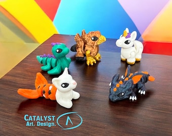 May include: A collection of colorful fantasy creature figurines, including a green dragon, a griffin, a unicorn, a white cat-like creature with an orange tail, and a black dragon with orange accents. The figurines are on a wooden surface. The text "CATALYST Art. Design." is visible.