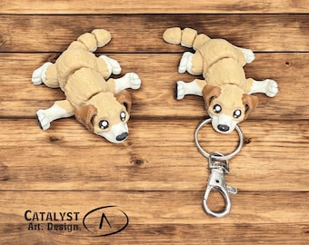May include: Two tan and white dog-shaped keychains and a matching figurine are displayed on a wooden surface. The keychains have a silver clasp and ring. The figurine and keychains have brown ears and black eyes. The text "CATALYST Art. Design." is visible.
