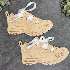 Könnte beinhalten: Zwei hölzerne schuhförmige Spielzeuge mit weißen Schnürsenkeln. Die Schuhe sind zum Üben des Schnürsens gedacht. Der Text "Color Me & Practice Shoe Tying" ist auf jedem Schuh aufgedruckt. Ein Schuh hat einen Stern und den Namen "Amara" aufgedruckt. Der andere Schuh hat ein Flammenmuster und den Namen "Logan" aufgedruckt.
