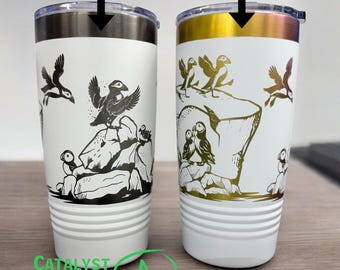 May include: Two stainless steel tumblers with a white background and a black and gold design of puffins on rocks. The tumbler on the left has a black design and the tumbler on the right has a gold design. The tumblers are labeled "Ghost Black" and "Rainbow".