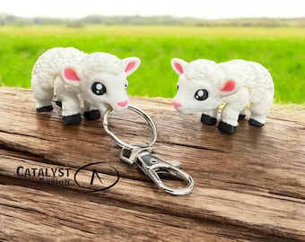 May include: A keychain featuring two white lamb figurines with pink ears and black hooves. The lambs are attached to a silver-colored keyring and clasp. The word "CATALYST" is visible on the wooden surface.