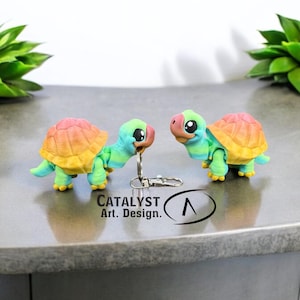 May include: Two small, colorful, 3D printed turtle keychains. The turtles are facing each other and have a rainbow gradient color scheme. The keychains are on a gray surface with the text "CATALYST Art. Design." below them.