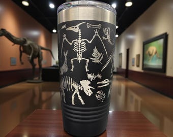 May include: Black travel tumbler with a silver lid, featuring etched white illustrations of dinosaur skeletons, ferns, and other prehistoric elements. The tumbler is cylindrical and sits on a wooden surface.
