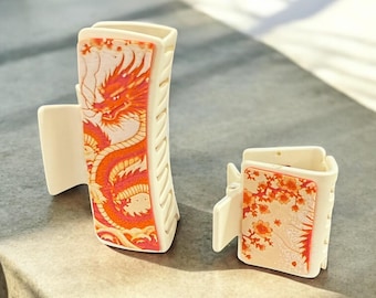 May include: Two white plastic hair clips with decorative designs. The larger clip features a red and orange dragon design. The smaller clip features a pink and white floral design. The text "CATALYST Art. Design." is visible in the bottom right corner of the image.
