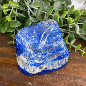 May include: A polished lapis lazuli stone with a deep blue color and white and gold veining. The stone is irregularly shaped and sits on a wooden surface, with green foliage in the background. A natural gemstone.