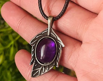 Op de afbeelding: Een zilveren ketting met een hanger met een paarse amethist edelsteen in een bladerenontwerp. De ketting heeft een bruin koord.