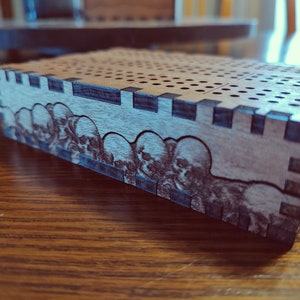 May include: A wooden box with a grid pattern on the top and a skull design on the side. The box is made of light brown wood with dark brown accents.