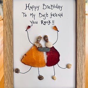 May include: A framed birthday card with a handwritten message that reads "Happy Birthday To My Best Friend You Rock!!". The card features a handmade art piece made with seashells and stones, depicting two figures holding hands.