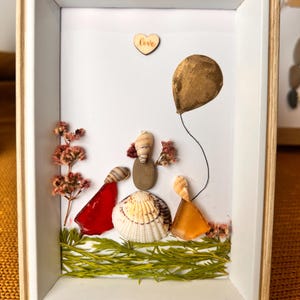 May include: Framed artwork depicting a family crafted from seashells and stones, with a gold-coloured balloon. The piece incorporates dried flowers and a wooden heart with the word "Love". The frame is white with a natural wood border. The artwork is in a rectangular frame.