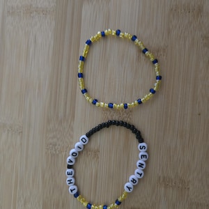 May include: Two beaded bracelets. One bracelet is yellow and blue. The other bracelet is black, yellow, and blue, with the words "THE VOID" and "SENTRY" spelled out in white letter beads.