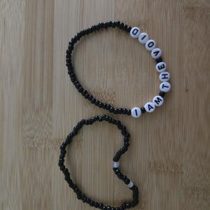 May include: Two beaded bracelets. One bracelet is black with white letter beads spelling "I AM THE VOID". The other bracelet is black with a few white beads. The bracelets are on a light brown wooden surface.