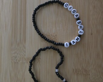May include: Two beaded bracelets. One bracelet is black with white letter beads spelling "I AM THE VOID". The other bracelet is black with a few white beads. The bracelets are on a light brown wooden surface.