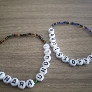 May include: Two beaded bracelets with white beads spelling out the names "Luminara" and "Barris Offee". The beads are strung on brown, black, and green thread for the first bracelet and blue, black, and yellow thread for the second bracelet.