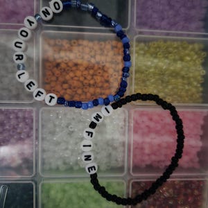 May include: Two beaded bracelets. One bracelet is white with black letters spelling "NO YOUR LEFT". The other bracelet is black with white letters spelling "I'M FINE". The bracelets are in a clear plastic organizer.