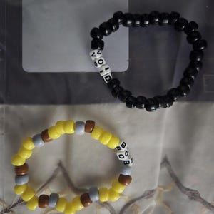 May include: Two beaded bracelets, one black with white beads spelling "VOID" and one with yellow, brown, and gray beads spelling "BOB".