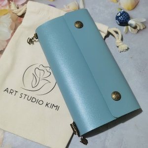Puede incluir: Un diario de cuero azul claro con dos cierres de botón de latón y una cadena de metal decorativa. El diario está sobre una bolsa de tela blanca con el texto "ART STUDIO KIMI" impreso en ella.
