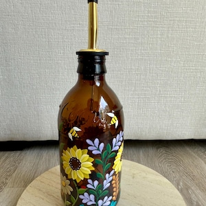May include: A brown glass bottle with a gold spout and black cap, decorated with hand-painted sunflowers, bees, and other flowers. The bottle is sitting on a wooden surface.