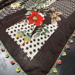 May include: A brown fabric square with a floral print and a crocheted border. The fabric features red, yellow, and green flowers on a white background. The crocheted border is green, yellow, and red.