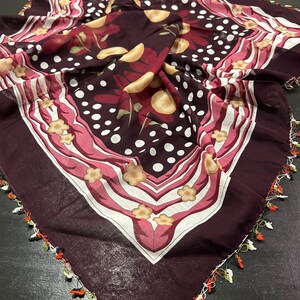 May include: A burgundy square scarf with a white, pink, and yellow floral pattern. The scarf has a crocheted border with white, green, orange, and black accents.