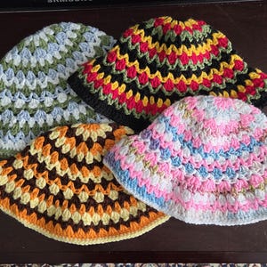 May include: Four handmade crochet bucket hats featuring granny square patterns. One hat has light blue, white, and green yarn. Another features black, red, yellow, and green. A third has orange, brown, and yellow. The fourth is pink, blue, and white.