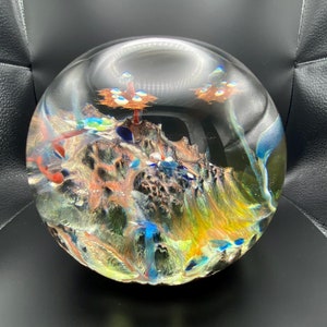 Puede incluir: Un pisapapeles de vidrio con un diseño abstracto colorido. El diseño presenta una variedad de colores, incluyendo azul, verde, naranja y rojo. El pisapapeles es redondo y tiene una superficie lisa y pulida.