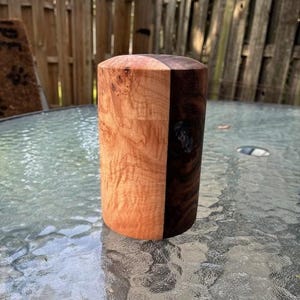 May include: A cylindrical wooden object, split vertically into two contrasting halves. One side displays a light, warm-toned wood with intricate grain patterns, while the other is a darker, richer brown. The top is flat, and the object stands on a glass surface.