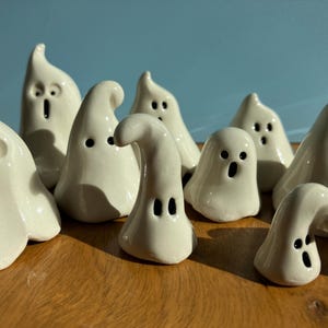 May include: A group of 10 white ceramic ghost figurines with black eyes and mouths. The ghosts are arranged in a semi-circle on a wooden surface.