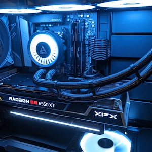 Puede incluir: Una vista cercana del interior de una caja de ordenador con un esquema de color azul y negro. La caja cuenta con un sistema de refrigeración líquida personalizado con tubos negros y un ventilador blanco. La tarjeta gráfica es una XFX Radeon RX 6950 XT.