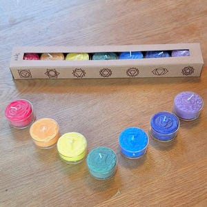 Op de afbeelding: Een set van zeven chakra theelichtjes in een kartonnen doos. De kaarsen zijn gerangschikt in een regenboogpatroon, met rode, oranje, gele, groene, blauwe, indigo en violette kaarsen. De doos heeft een label met de zeven chakra symbolen.
