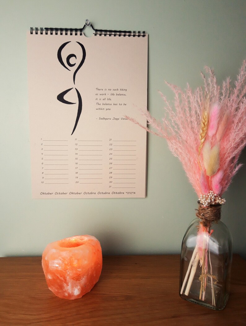 May include: A pink and white dried flower arrangement in a clear glass vase with a brown twine accent. The vase is on a wooden table with a salt lamp next to it. A calendar with the quote "There is no such thing as work-life balance, it is all life. The balance has to be within you. - Sadhguru Jaggi Vasu" is hanging on a wall behind the vase.