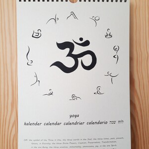 May include: A white calendar with a black Om symbol in the center. The calendar is titled "yoga" and has the words "kalender, calendar, calendrier, calendario, לוח שנה" written below the title.