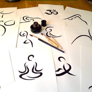 May include: A collection of white paper sheets with black ink drawings of yoga poses and the Om symbol. Two paintbrushes and a bottle of black ink are on the table.