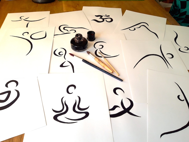 May include: A collection of white paper sheets with black ink drawings of yoga poses and the Om symbol. Two paintbrushes and a bottle of black ink are on the table.