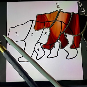 May include: A stained glass bear with brown and black glass pieces. The bear is hanging from a chain. A white outline of a bear with numbers 1 through 5 is on a white surface. A glass cutter and a stylus are also on the surface.