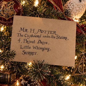 May include: A rectangular tan envelope with black calligraphy text reading: Mr H. Potter, The Cupboard under the Stairs, 4, Privet Drive, Little Whinging, Surrey. The envelope is positioned against a background of green pine needles and lights.