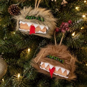 May include: Two fuzzy monster ornaments hanging by twine. One is light tan and the other is medium brown. Each features four green bead eyes, a row of white felt teeth, and a red felt tongue protruding from a wooden block mouth structure.