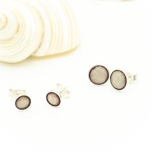 Könnte beinhalten: Silberne Ohrstecker mit einem kreisförmigen, hellgrauen Mittelteil und einem dunkelgrauen Rand. Die Ohrringe werden auf weißem Hintergrund mit zwei Muscheln präsentiert.