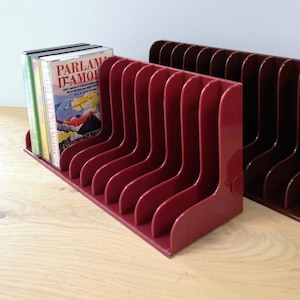 May include: Two red plastic cassette tape holders. The holder on the left has a stack of cassette tapes in it. The holder on the right is empty.