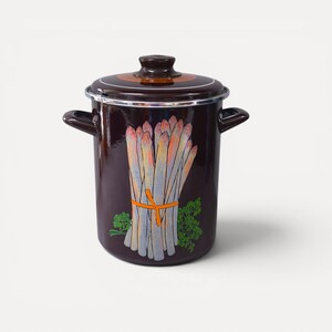 Può includere: Una pentola in smalto marrone scuro con coperchio abbinato e due manici laterali. La parte anteriore della pentola presenta un'illustrazione grafica di un mazzo di asparagi bianchi legati con un nastro arancione e rametti di prezzemolo verde.