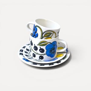 Op de afbeelding: Twee witte keramische kopjes gestapeld op een bijpassende schotel, versierd met een botanisch patroon van grote blauwe bloemen, gele bladeren en donkerblauwe bessen. Het ontwerp loopt rond de buitenkant van de kopjes en de rand van de schotel.