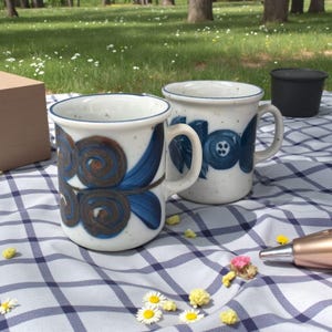 Op de afbeelding: Twee keramische mokken met blauwe en bruine wervel- en bloemmotieven. De mokken hebben witte achtergronden en blauwe randen. Ze staan op een blauw-wit geruite deken, met een groene achtergrond van gras en bomen.